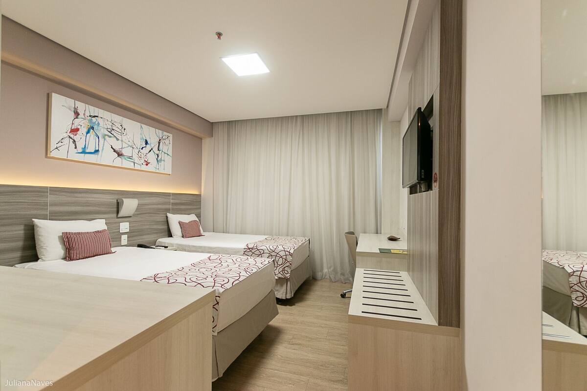 Habitación de hotel con dos camas, un escritorio, un televisor y un ventanal grande con cortinas.