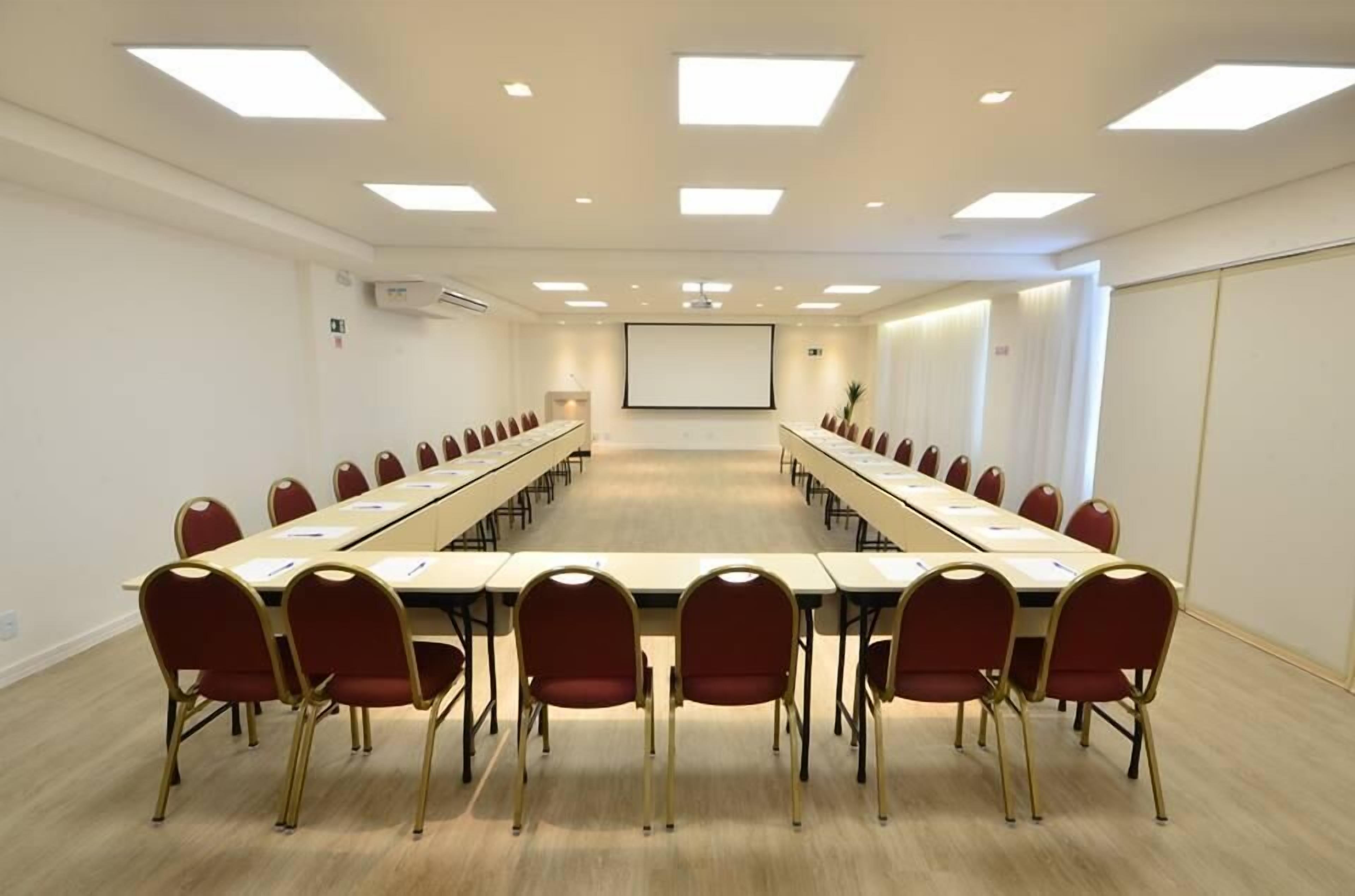 A conference room with a long table, red chairs, and a projector screen.