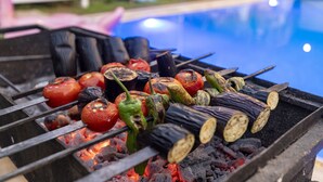 Grill med forskellige grøntsager og kødspyd over kul, med en pool i baggrunden.