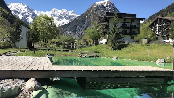 Eine Holzterrasse über einem klaren Pool, mit einem Berg im Hintergrund.
