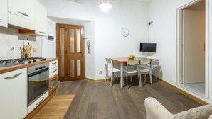 Une cuisine compacte avec des meubles blancs, un plan de travail en bois et un four encastré. On y trouve également un coin repas avec une table et des chaises, ainsi qu’un téléviseur fixé au mur.