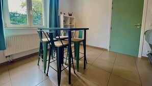 A room with a dining table and chairs, a radiator, a window with curtains, a green door, and a white shelf unit. - L'appartement d'Houyet (Houyet)