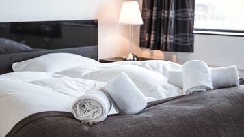 A neatly made bed with white linens and rolled towels, a bedside lamp, and a window with curtains.