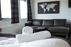 A neatly made bed with a rolled white towel, a dark leather sofa, a round coffee table, and a world map on the wall.