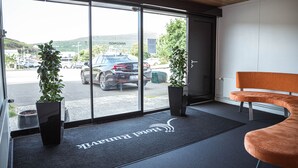 A hotel lobby with a reception desk, a bench, potted plants, and a view of the parking area through large glass doors. - Hotel Runavik (Runavik)