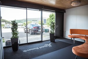 Eine Hotellobby mit einer Rezeption, einer Bank, Topfpflanzen und Blick auf den Parkplatz durch große Glastüren.