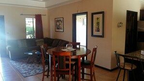 Uma área de refeições com uma mesa de madeira redonda, quatro cadeiras e uma tigela de vidro sobre a mesa. Há um sofá e uma janela com cortinas ao fundo.