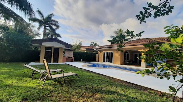 A backyard with a swimming pool, a lounge chair, and a small building with a door and windows.