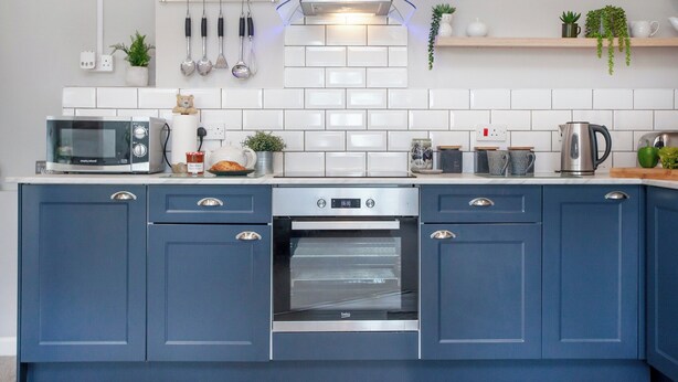 A modern kitchen with a built-in oven, microwave, and kettle. There are cabinets with handles, a countertop with various kitchen items, and a hanging rack with utensils.