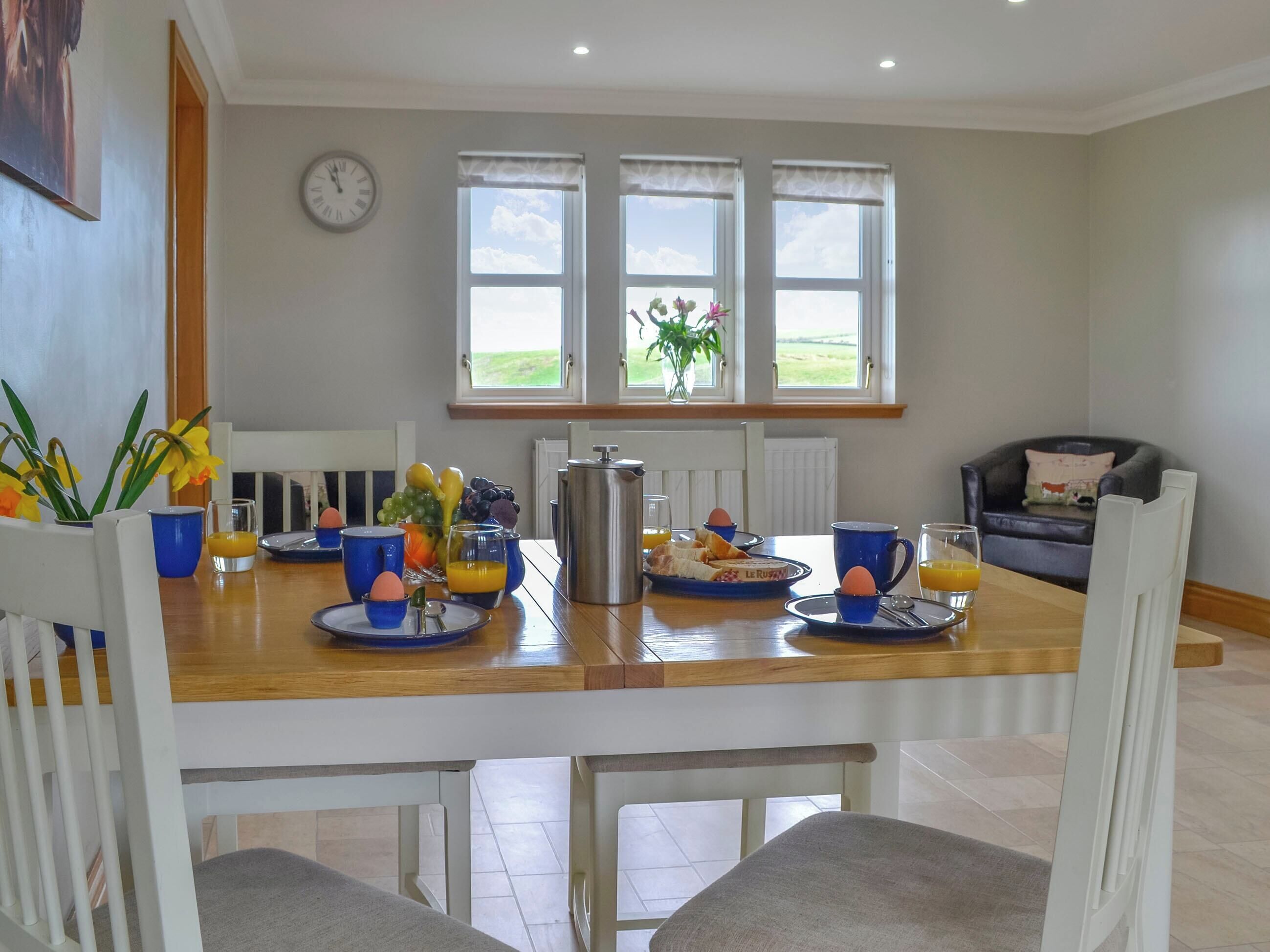 Uma sala de jantar com mesa de madeira, xícaras azuis e itens de café da manhã. Há janelas com cortinas, um relógio na parede e vista para o exterior.