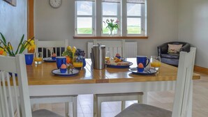 A dining area with a wooden table, blue cups, and breakfast items. There are windows with curtains, a clock on the wall, and a view of the outdoors. - 4 bedroom accommodation in Portpatrick, near Stranraer (Portpatrick, near Stranraer)