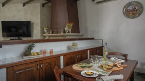 A kitchen with wooden cabinets, a dining table set with food and drinks, a TV on the wall, and a decorative plate on the wall. - Villa Alessia 6, Emma Villas (Gela)