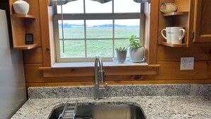 Una cocina con encimera de granito, pileta de acero inoxidable, armarios de madera, una ventana con vista a un campo y una planta en maceta en el alféizar.