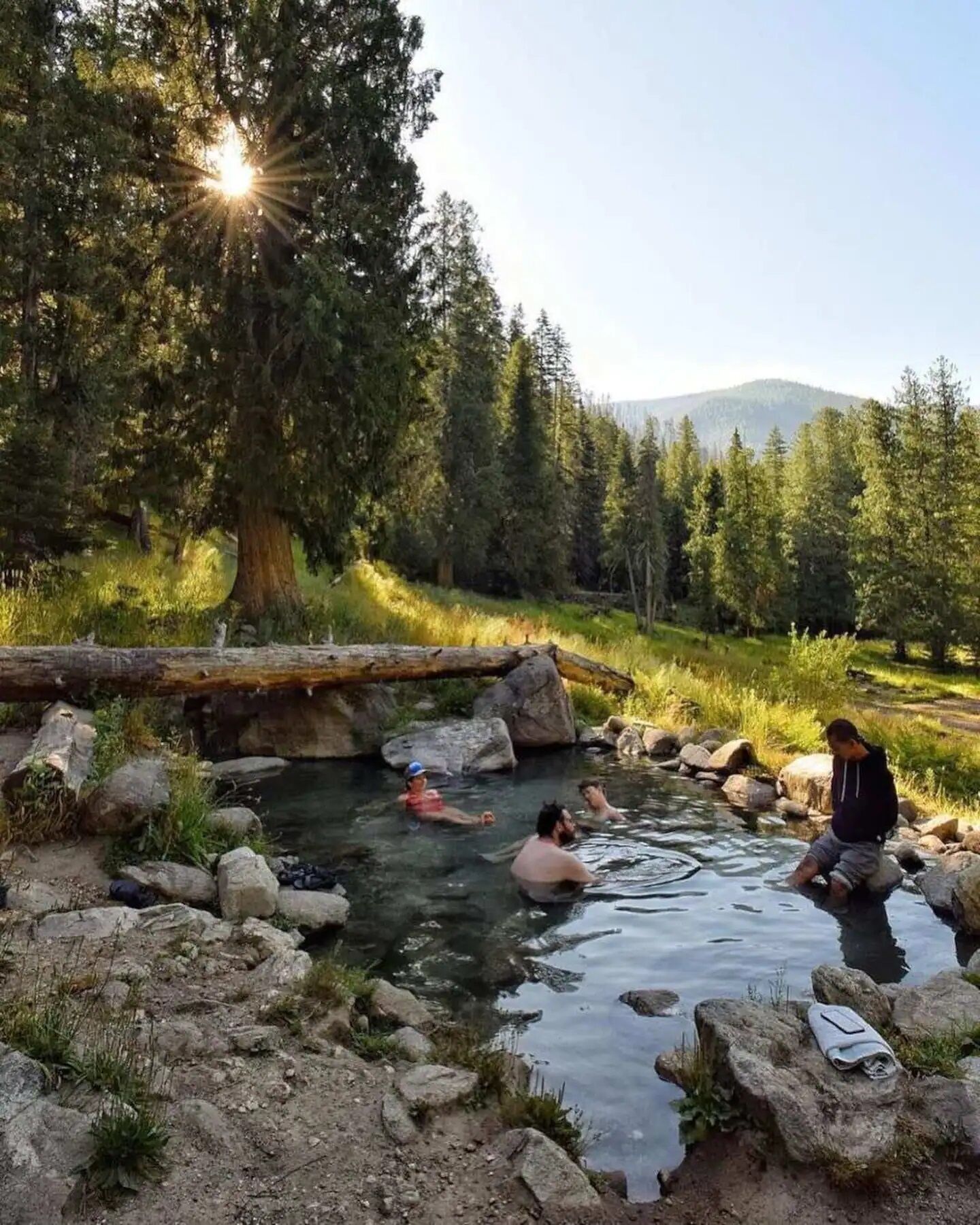 A natural hot spring with three people in the water, surrounded by trees and rocks.