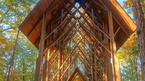 Een houten paviljoen met een puntdak, omgeven door een stenen pad en een dicht bos.