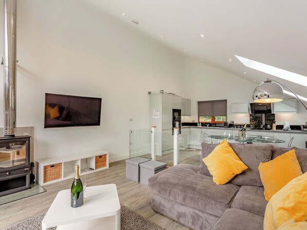 A modern living room with a sofa, a TV, a glass table with a bottle, and a kitchen in the background.
