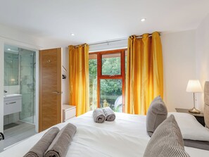 A hotel room with a bed, a bathroom with a glass shower, a window with curtains, and a nightstand with a lamp.