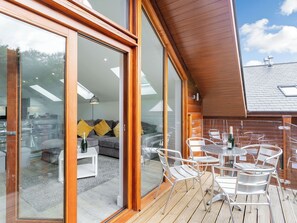 Um deck de madeira com área de estar externa e uma porta de vidro que dá para um espaço interno com sofá e mesa.