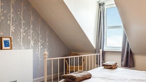 Ein Schlafzimmer mit schrägen Wänden, einem Bett mit gemustertem Bettbezug, einem Fenster mit Vorhängen und einer Wand mit Tapete mit Zweigen.