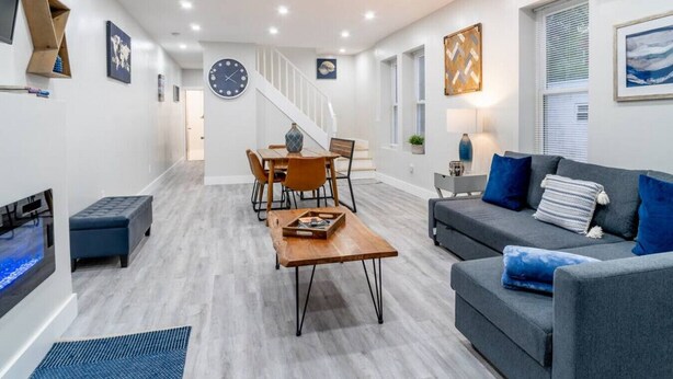 Una sala de estar moderna con un sofá gris, cojines azules, una mesa de centro de madera y un comedor con una mesa y sillas de madera.