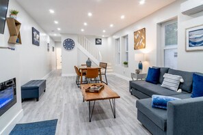 Uma sala de estar moderna com um sofá cinza, almofadas azuis, uma mesa de centro de madeira e uma área de jantar com uma mesa e cadeiras de madeira.