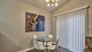 A dining area with a round glass table and four chairs, a ceiling light fixture, a large window with blinds, and a piece of wall art. - Large Groups Welcome (Go Seminoles, Go Rattlers) (Tallahassee)