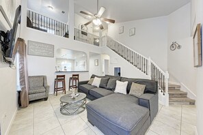 Ein geräumiges Wohnzimmer mit einer grauen Eckcouch, zwei Holzstühlen, einem runden Couchtisch und einer Treppe mit weißer Geländer, die zu einer oberen Ebene führt.