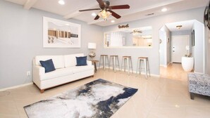 Una sala de estar moderna con un sofá blanco, un ventilador de techo, una zona de bar y una cocina visible a través de una puerta abierta.