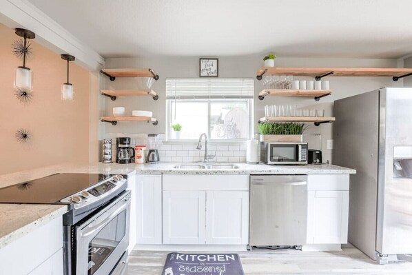 A modern kitchen with white cabinets, a stainless steel refrigerator, a microwave, and a coffee maker. There are wooden shelves above the sink and a window with blinds.
