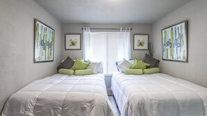 Une chambre avec deux lits, du linge de lit blanc, des oreillers verts et gris, et des œuvres d’art encadrées accrochées aux murs.