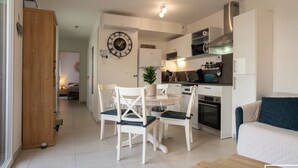 A modern kitchen with white cabinetry, a dining area with a round table and white chairs, and a living area with a sofa and a blue cushion.