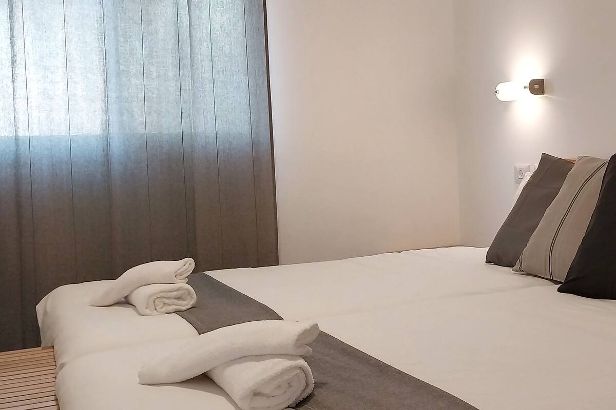 A hotel room with a neatly made bed, a grey throw blanket, and a wall-mounted light fixture.