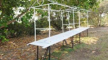 A white metal pergola structure with a bench underneath, surrounded by trees and fallen leaves.