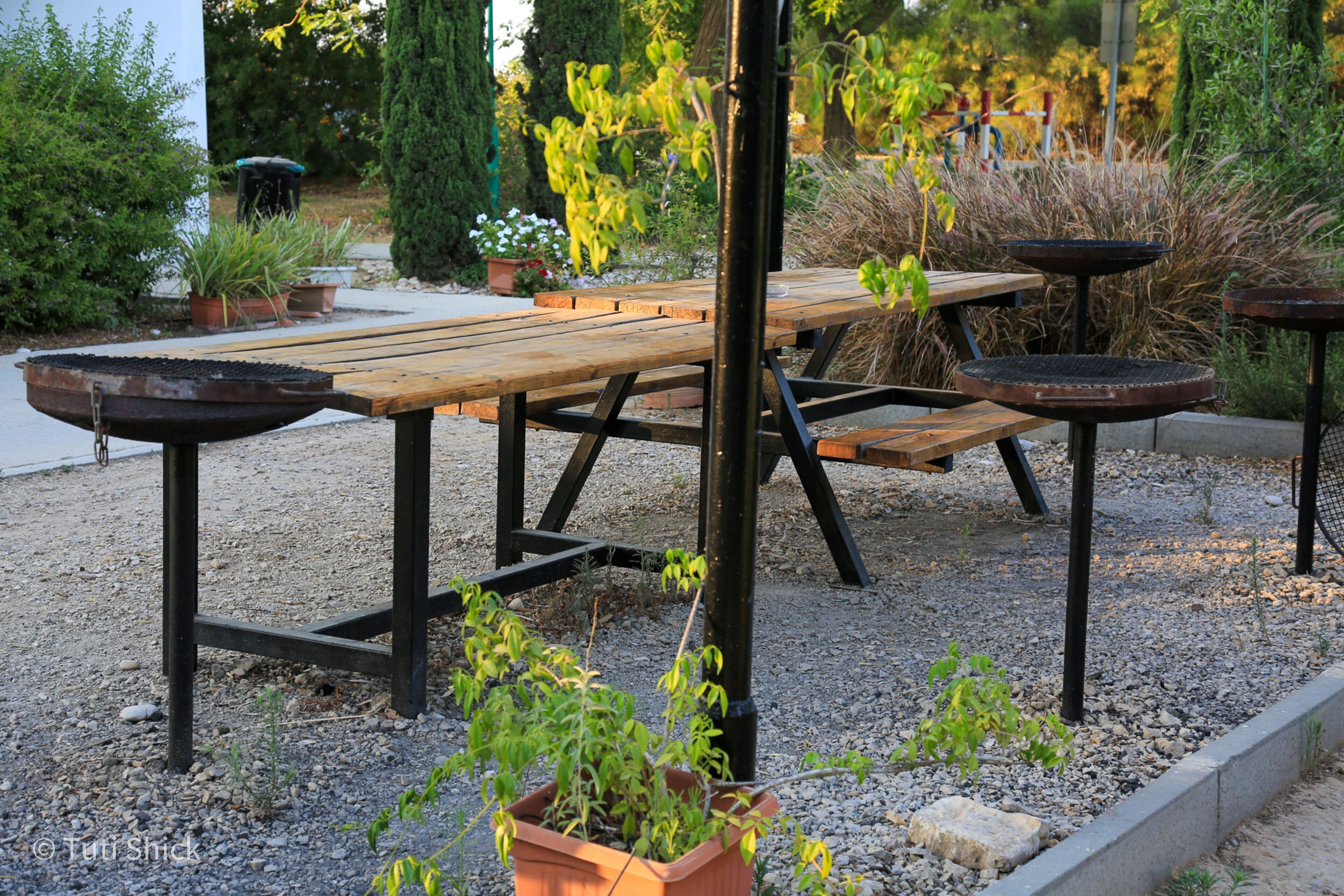 Une table de pique-nique en bois, dotée de pieds en métal, entourée de gravier et de plantes en pot.