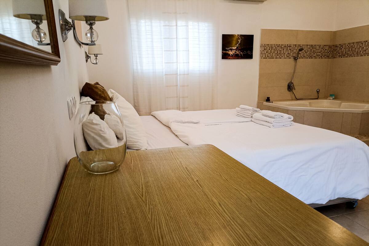 A hotel room with a wooden table, a bed with white linens, a glass vase with a bouquet, a bathroom with a bathtub, and a wall-mounted picture.