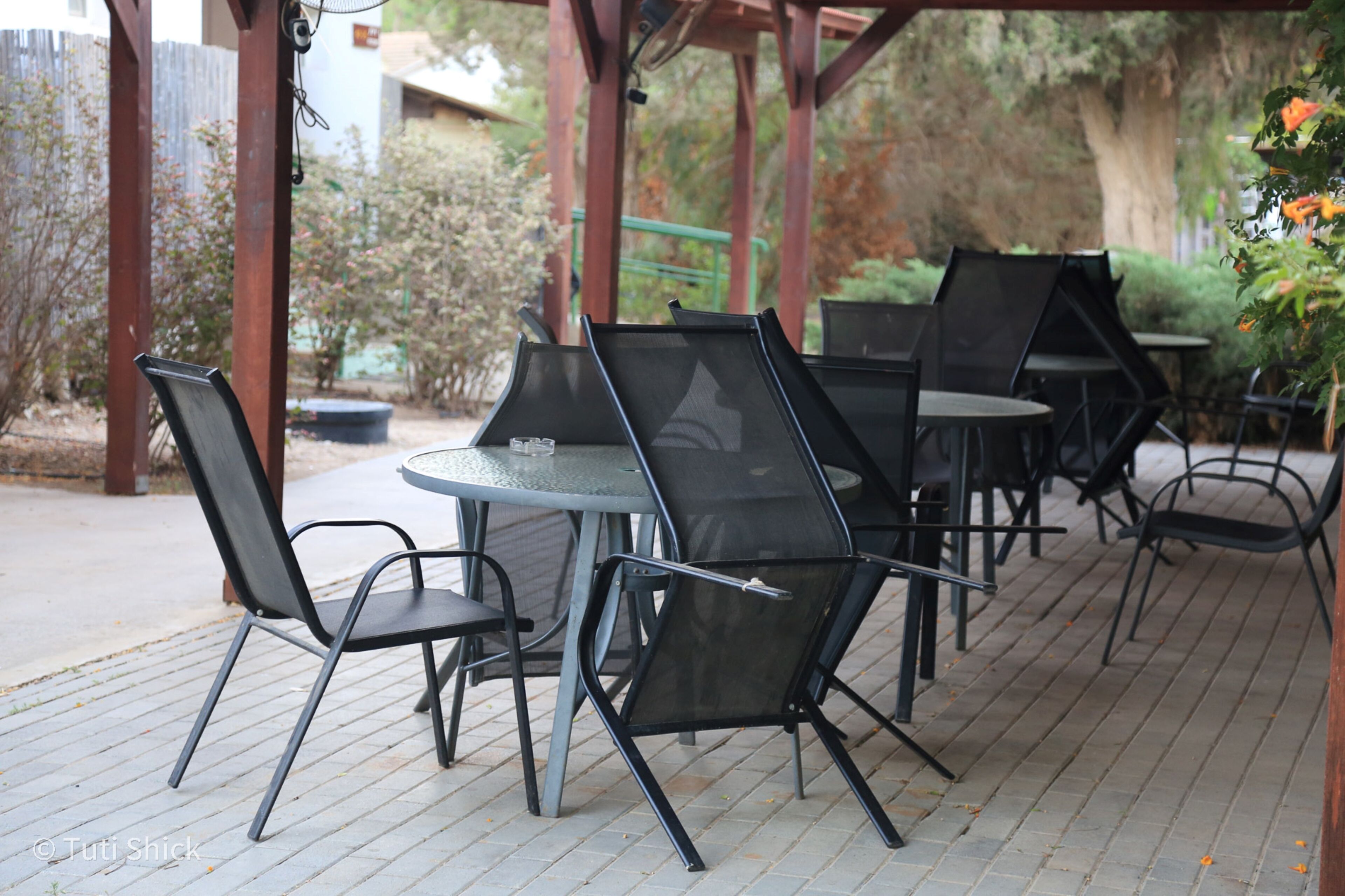 An outdoor patio with metal chairs and tables, surrounded by greenery and a wooden pergola.