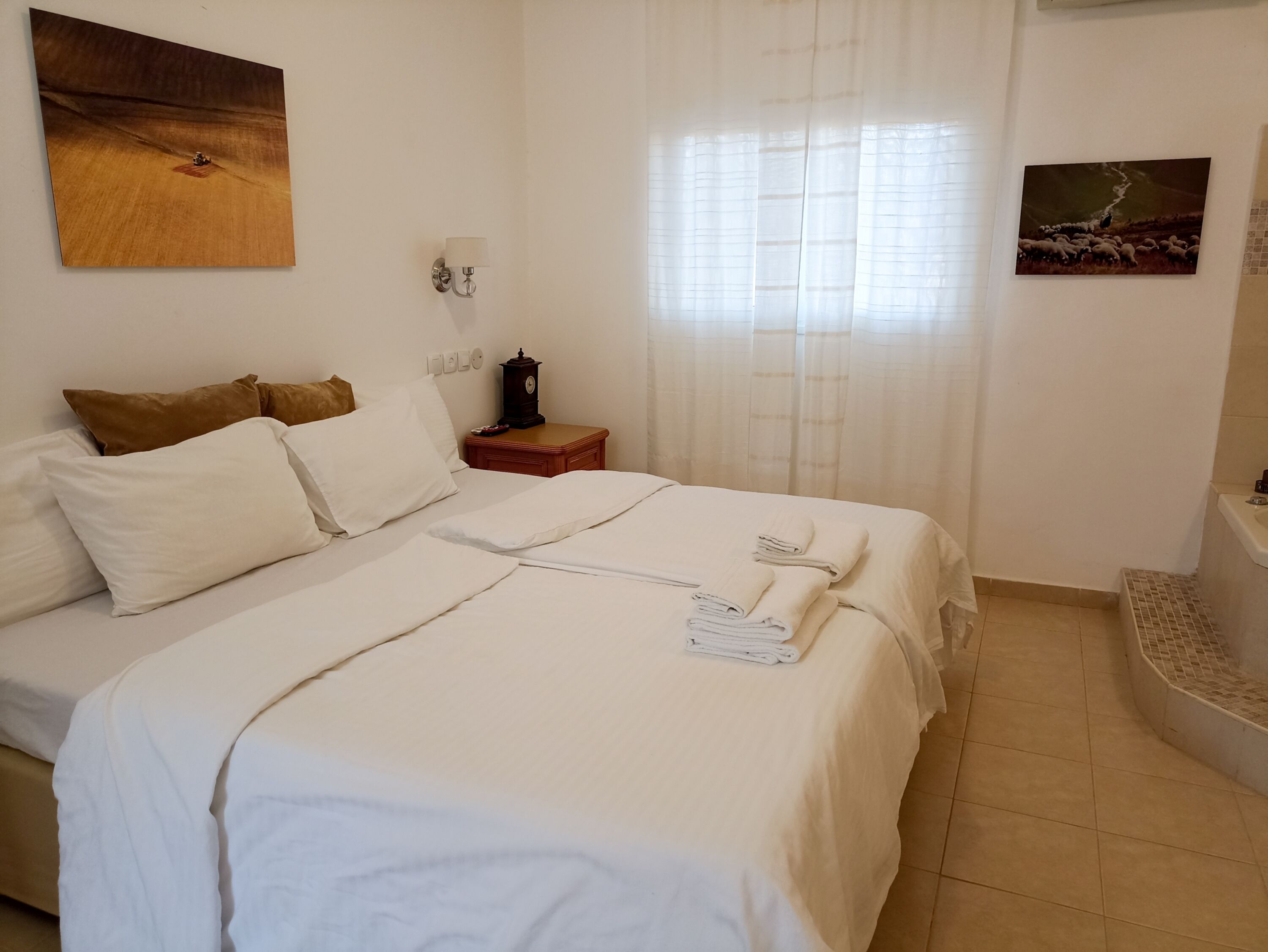 A neatly made bed with white linens, a bedside table with a clock, and a wall-mounted lamp.