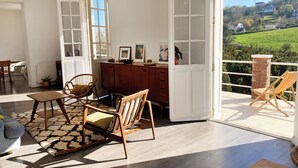 A spacious room with a wooden floor, a dining area with a table and chairs, a large window with a view of a green landscape, and a balcony with a chair. - Seaside villa in Normandy, exceptional setting (Saint-Pierre-en-Port)