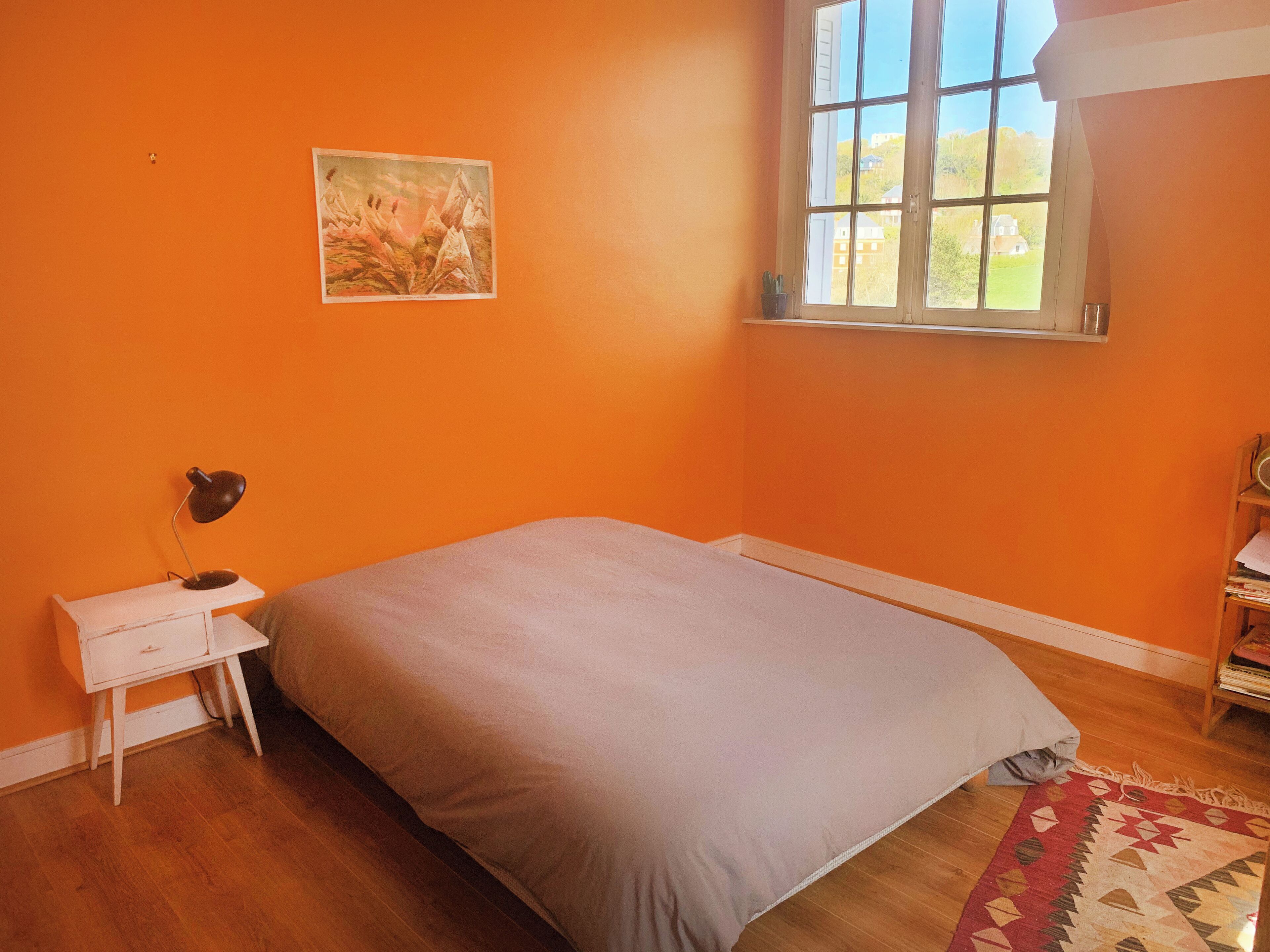 A bedroom with an orange wall, a bed with a grey bedspread, a small white nightstand with a lamp, a window with a view of greenery, and a patterned rug on the floor.