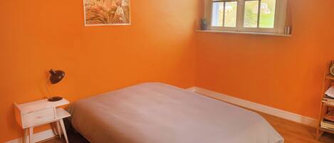 A bedroom with an orange wall, a bed with a grey bedspread, a small white nightstand with a lamp, a window with a view of greenery, and a patterned rug on the floor.
