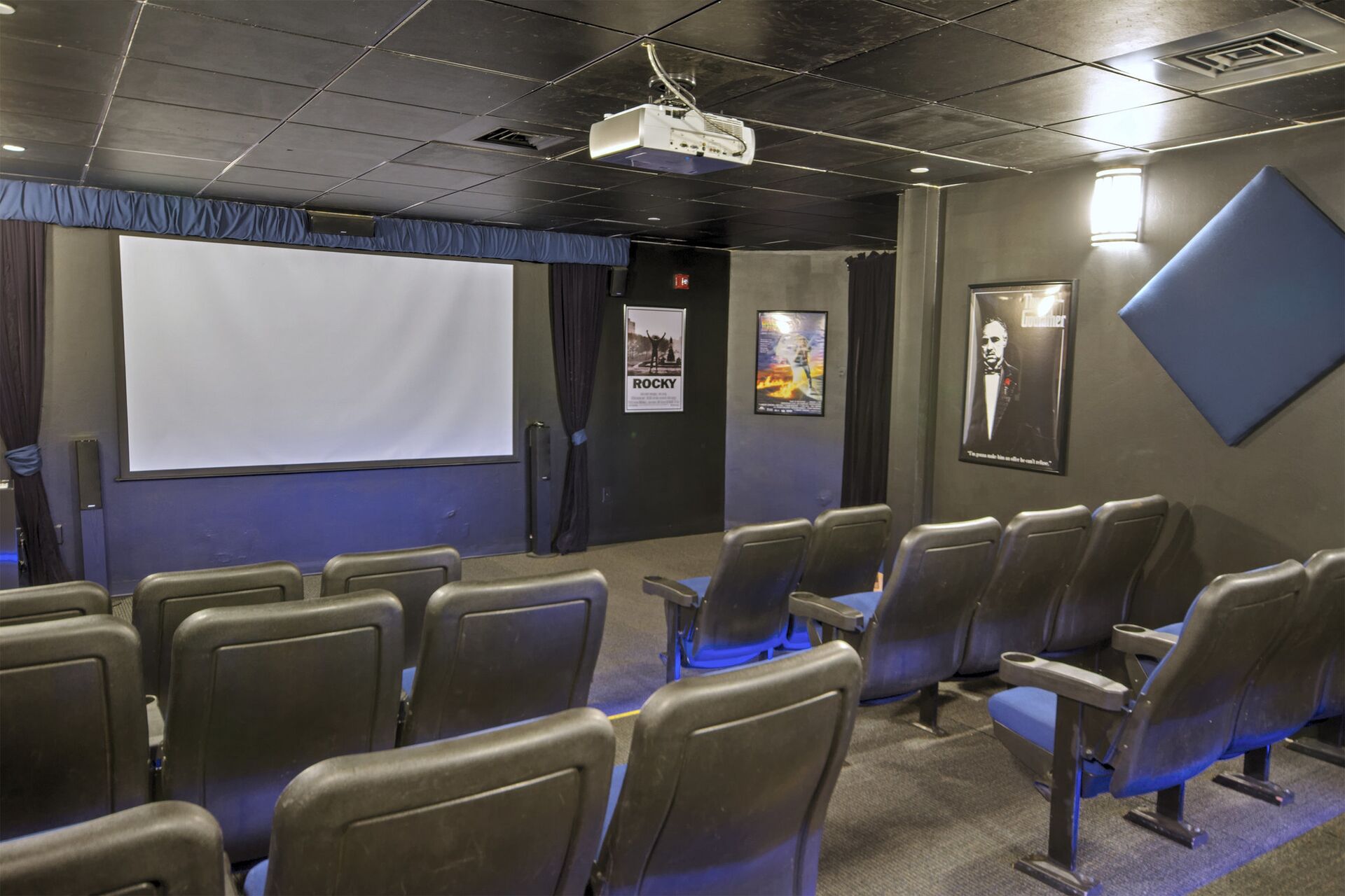 A small, dimly lit movie theater with rows of plain, grey seats and a large screen.