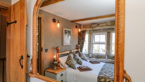 Un dormitorio con un espejo de madera, una cama con ropa de cama estampada, una mesita de noche con una lámpara y una pared de piedra.
