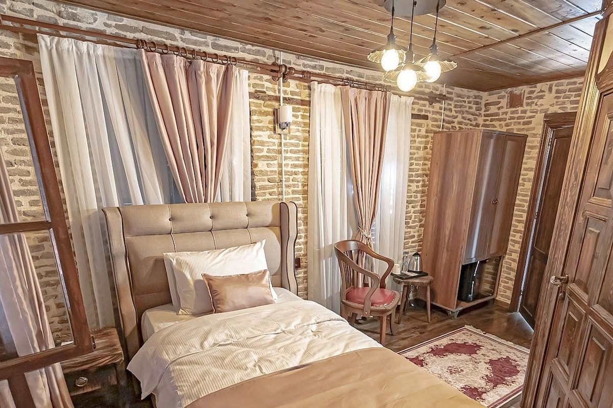Un dormitorio con una cama grande, cabecero de madera y pared de piedra. Hay una silla y una mesita de noche con una lámpara.