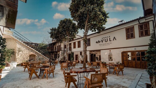 Ein Innenhof mit Außenbestuhlung, einer Treppe und einem Gebäude, das ein Schild mit der Aufschrift 'KONAK GAVULA BOUTIQUE HOTEL EST. 2020 ANTAKYA' zeigt.