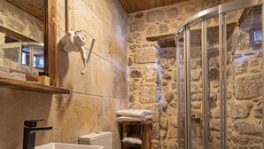 Ein Badezimmer mit Steinwand, einem Holzanrichte, einem weißen Waschbecken, einer Toilette und einer Duschkabine aus Glas.