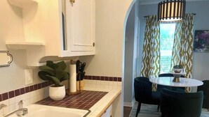 A kitchen with a sink, cabinets, and a potted plant. There is a dining area with a table and chairs, and a patterned curtain. - Lower Greenville Studio (Dallas)