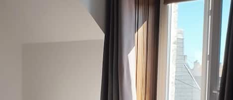 A bedroom with a bed, a nightstand, a window with curtains, and a view of buildings outside.