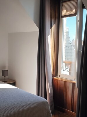 Un dormitorio con cama, mesita de noche, ventana con cortinas y vista a edificios.