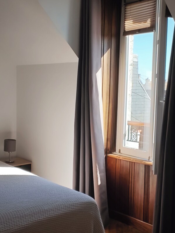 A bedroom with a bed, a nightstand, a window with curtains, and a view of buildings outside.