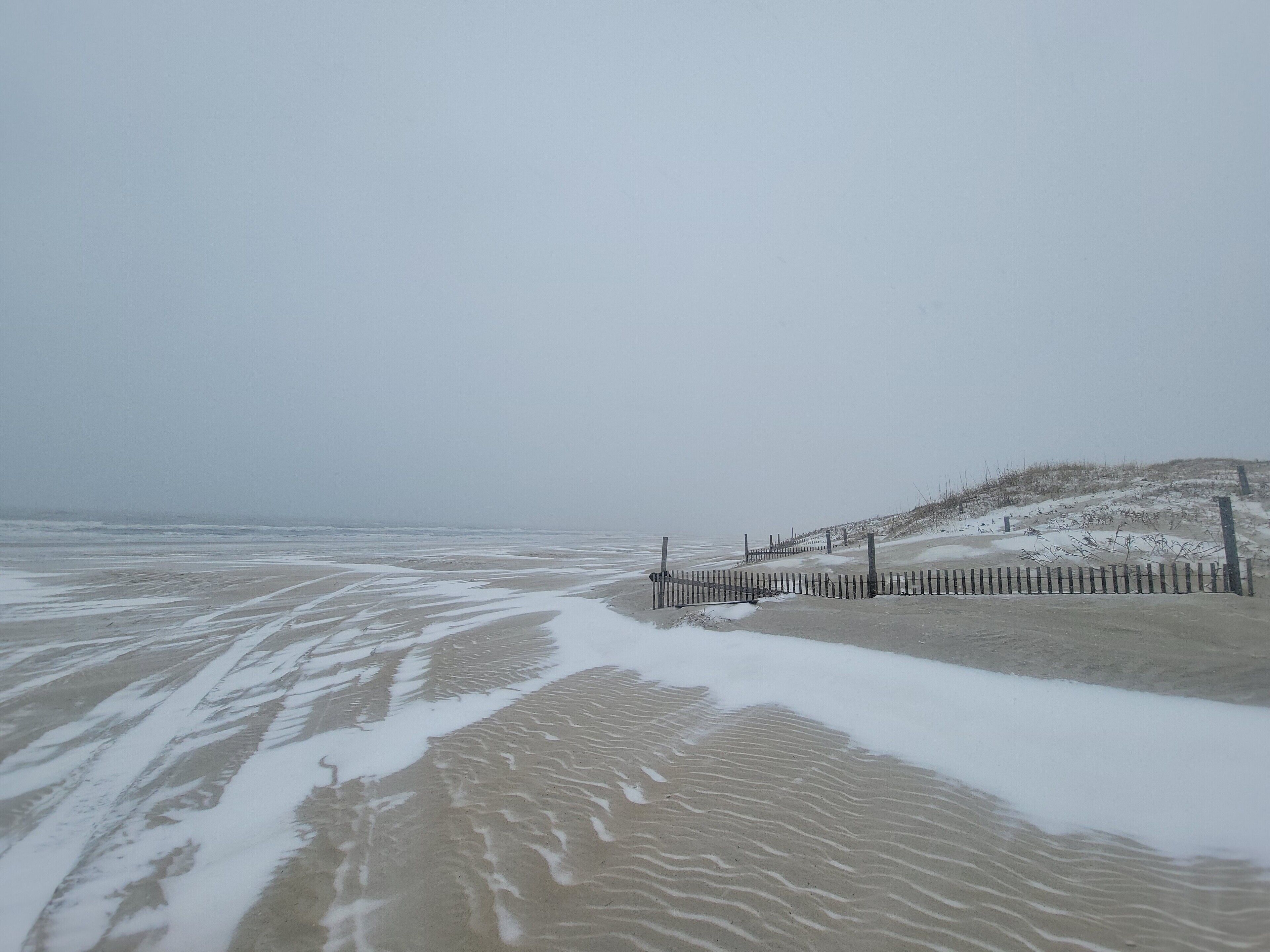 A beach with a wooden fence and snow on the sand.